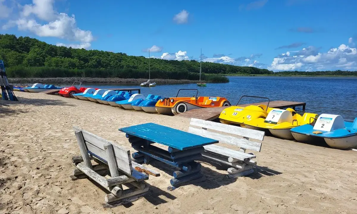 Słoneczny dzień nad jeziorem Sarbsko. Rząd kolorowych rowerów wodnych czeka na plaży, obok drewniane ławki i stół.