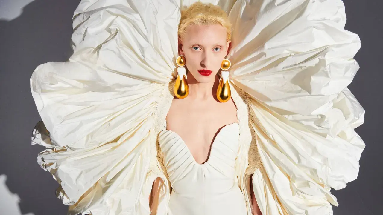 A model wears a dramatic white gown with a voluminous, ruffled cape, showcasing Daniel Roseberry's avant-garde design and striking gold teardrop earrings.