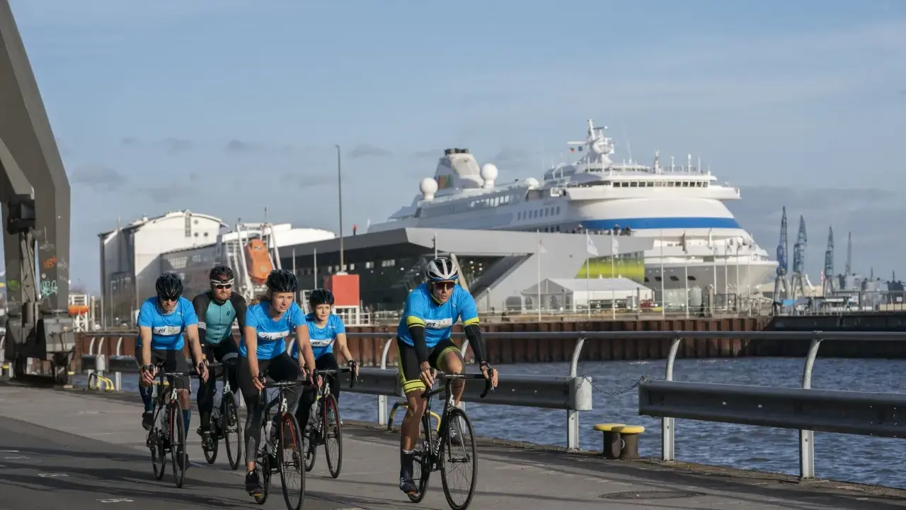 Fünf Radfahrer in blauen Trikots trainieren für einen Mini-Triathlon am Hafen, ein großes Kreuzfahrtschiff im Hintergrund.