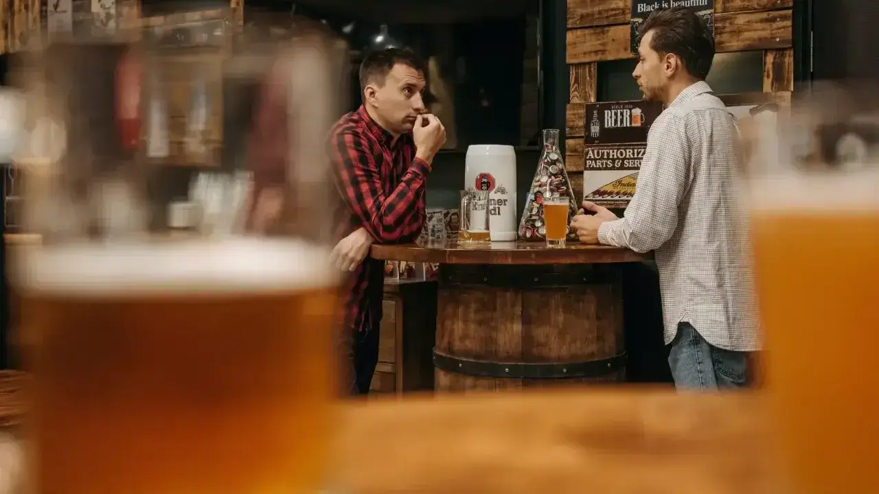 Dwóch mężczyzn rozmawia przy barze, zastanawiając się, ile gram alkoholu ma piwo. Na blacie stoją kufle i piwo.