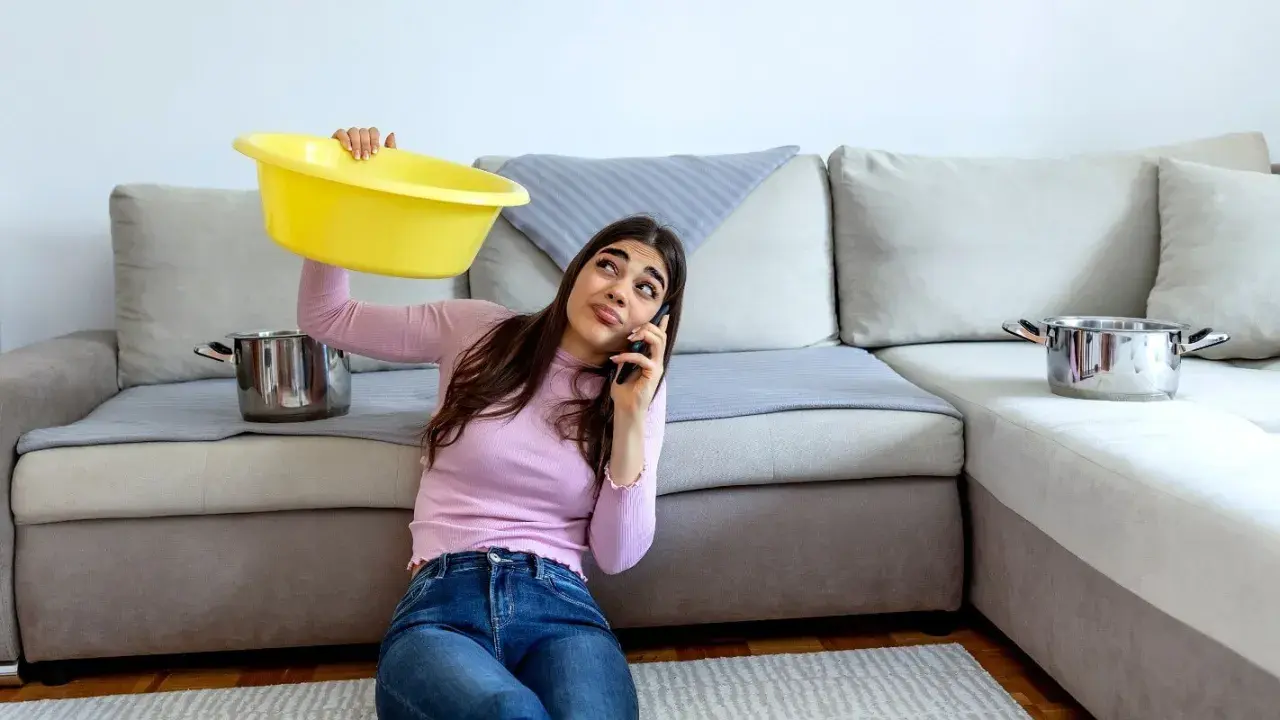 Mujer preocupada llama por teléfono mientras sostiene un barreño amarillo para recoger agua de las goteras en casa. Ollas esperan en el sofá.