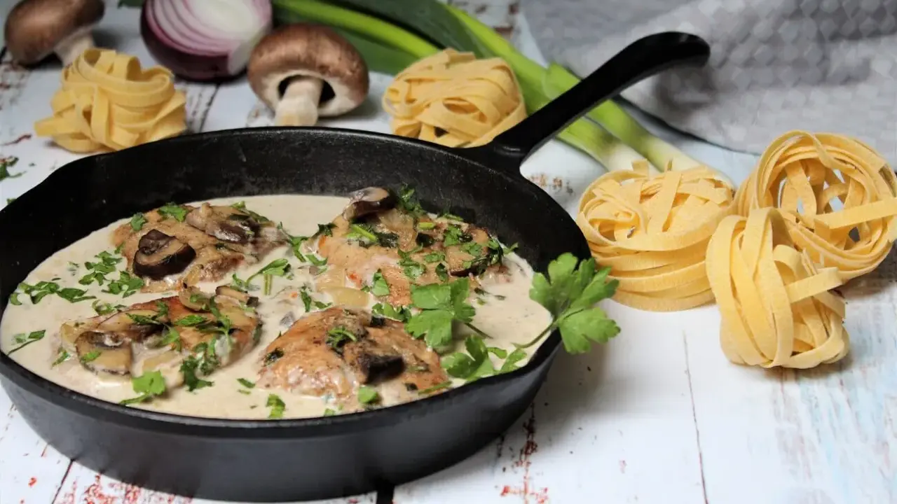 Ein köstliches Schnitzelpfanne Rezept mit cremiger Pilzsoße in einer Gusseisenpfanne, garniert mit Petersilie. Daneben liegen frische Pasta-Nester, Pilze und Lauch.