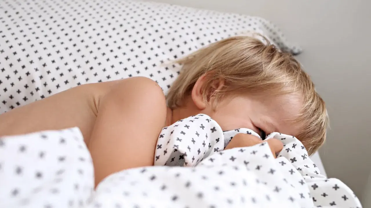 Niño rubio se esconde bajo la sábana. Aprende como hacer que un niño duerma solo con paciencia y rutinas.