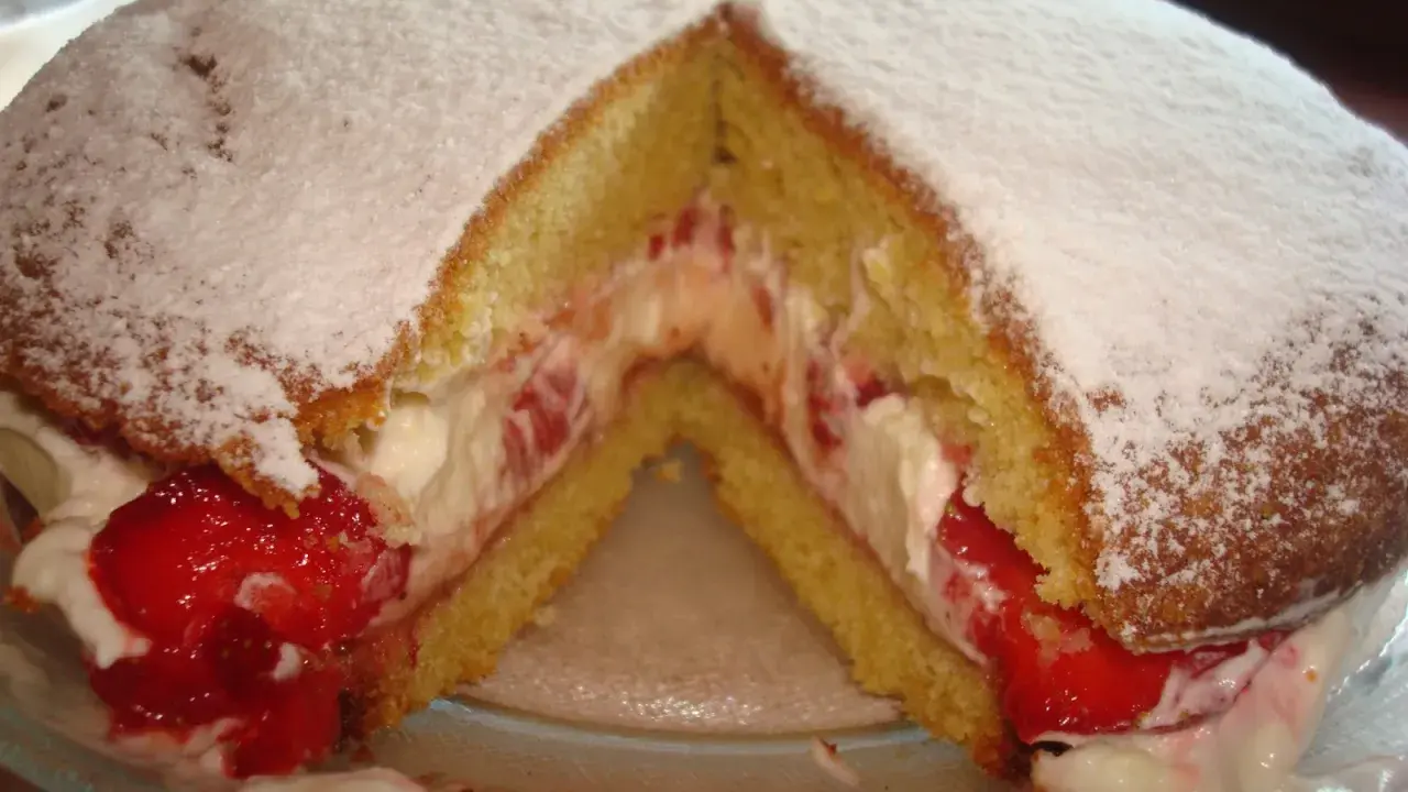 Puszysty biszkopt przełożony kremem i truskawkami, posypany cukrem pudrem. Klasyczny victoria sponge cake.