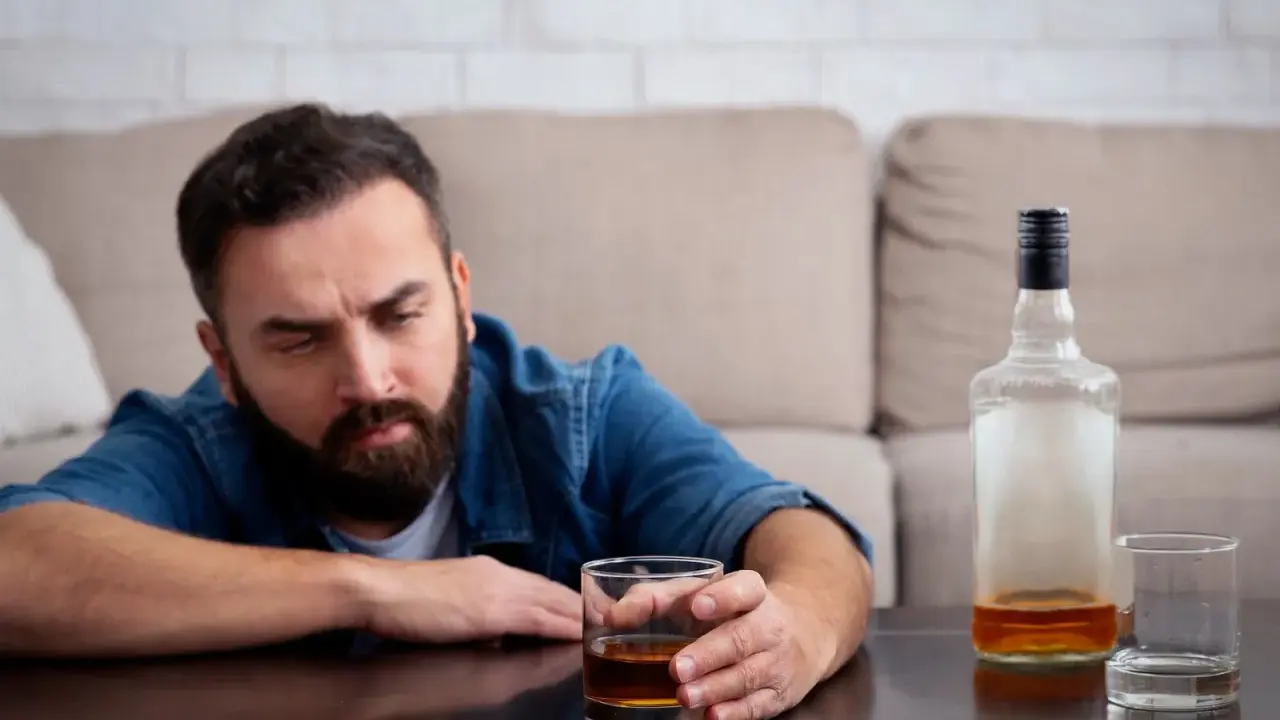 Mężczyzna z zarostem patrzy na szklankę z alkoholem, widoczne objawy uzależnienia. Obok pusta butelka i kolejna szklanka.