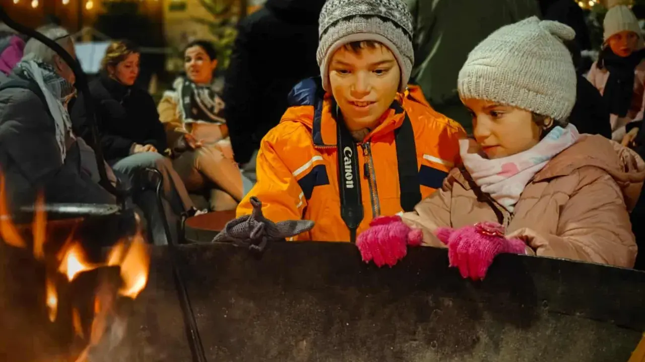 Niños disfrutan de una fogata en Italia, con luces festivas de fondo.