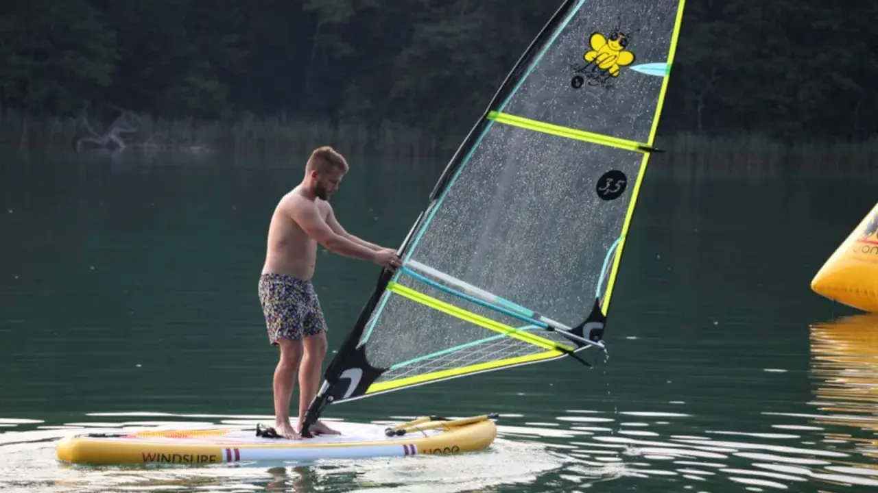 Mężczyzna na desce z żaglem, gotowy do wind sup. Na żaglu pszczółka i napis "3.5".