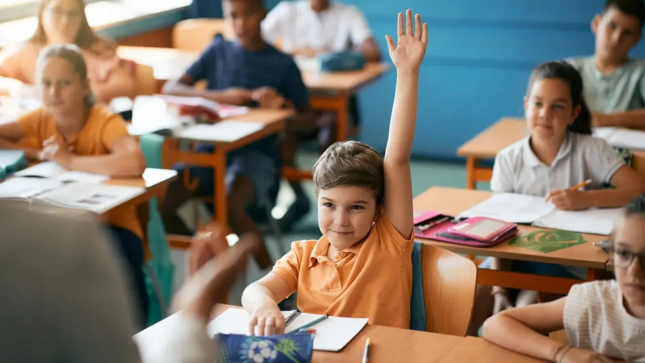 Chłopiec z podniesioną ręką czeka na ocenianie odpowiedzi. Inne dzieci pracują przy ławkach.