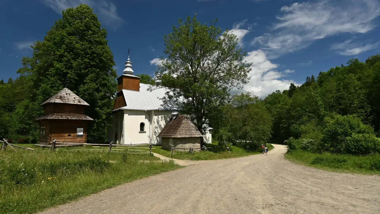 Cerkiew w Łopience, drewniana chata i kamienna przybudówka na tle zieleni i błękitnego nieba z chmurami.