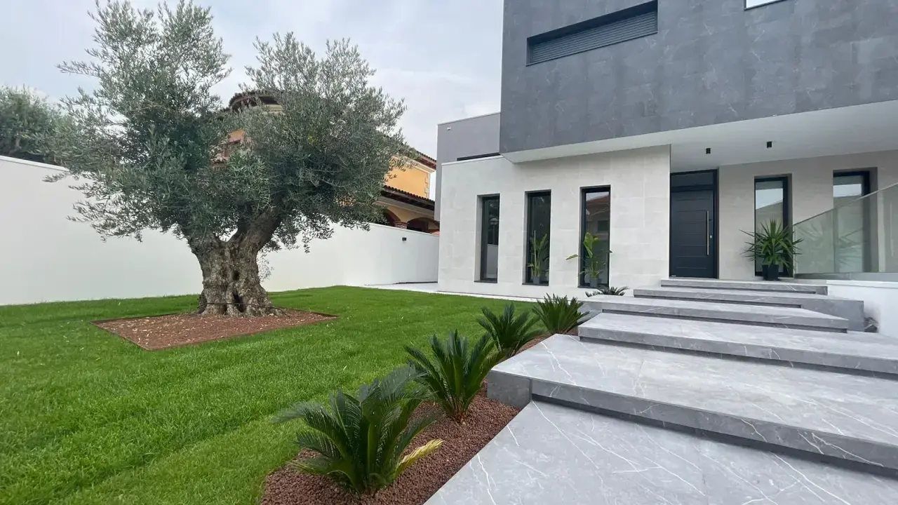 Moderna casa con fachadas color gris y blanco, escaleras de mármol y un olivo centenario en el jardín.