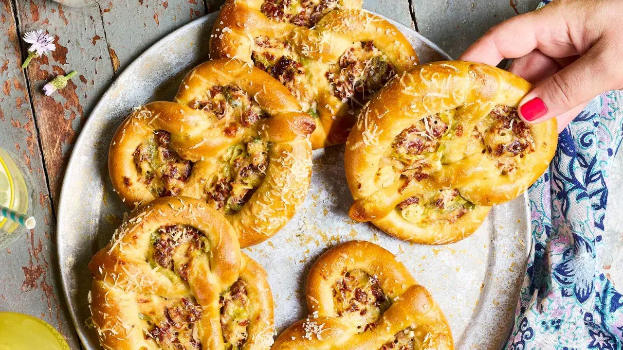 Herzhafte Brezeln mit Lauch und Speck, die an Flammkuchen erinnern, liegen auf einem Blech. Eine Hand greift nach einer Brezel.
