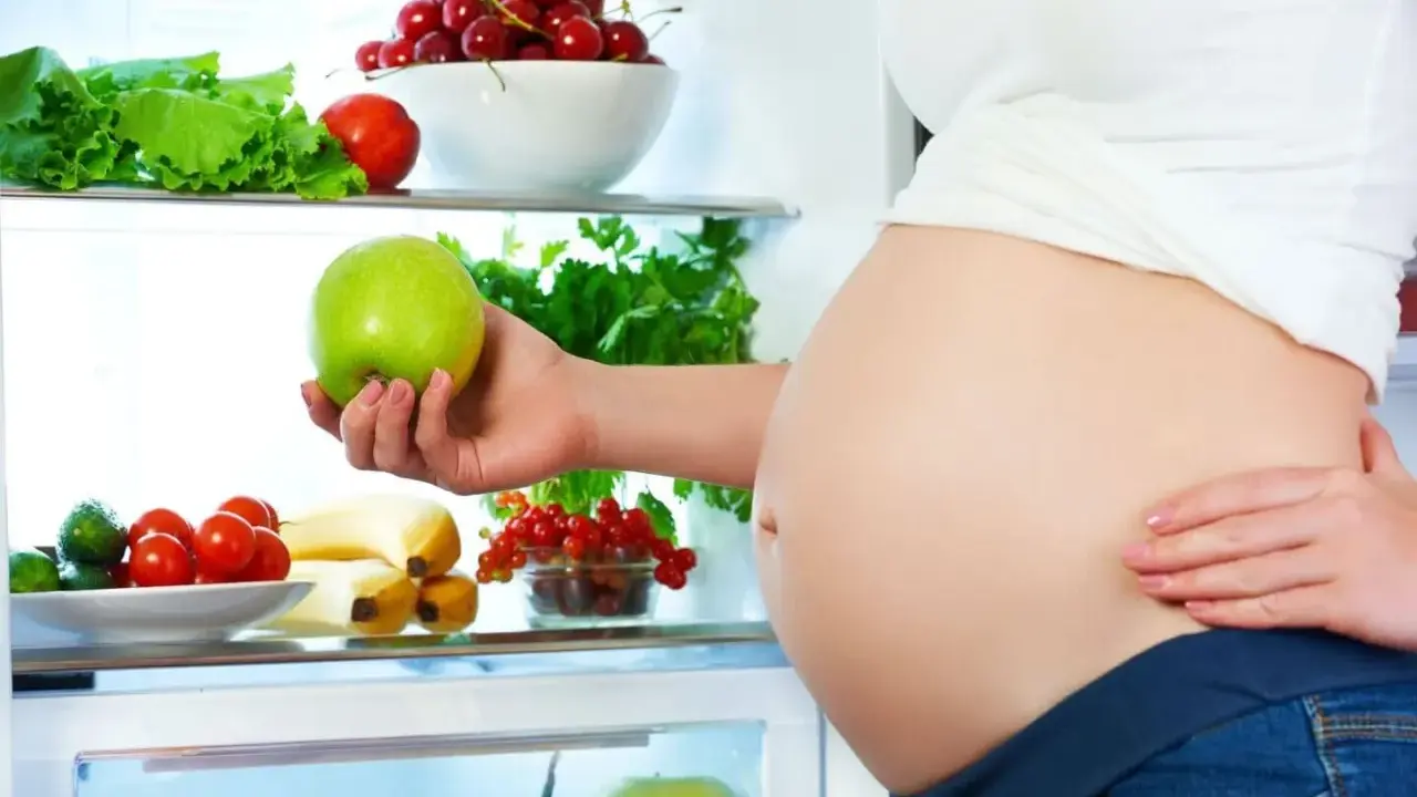 Gravid kvinna tar ett grönt äpple ur kylen fylld med färsk frukt och grönsaker. Hon njuter av hälsosam mat under sin soja graviditet.
