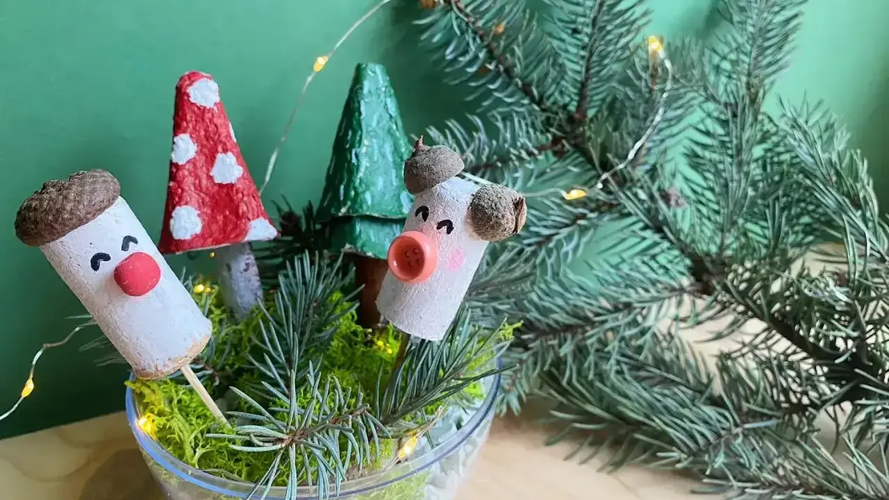 Uroczy stroik DIY z korkowych ludzików, grzybków i choinek w szklanej misie, ozdobiony gałązkami świerku i lampkami.