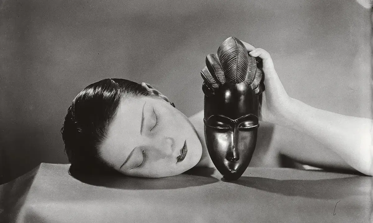 Foto Man Ray: volto femminile addormentato accanto a una maschera africana tenuta da una mano.