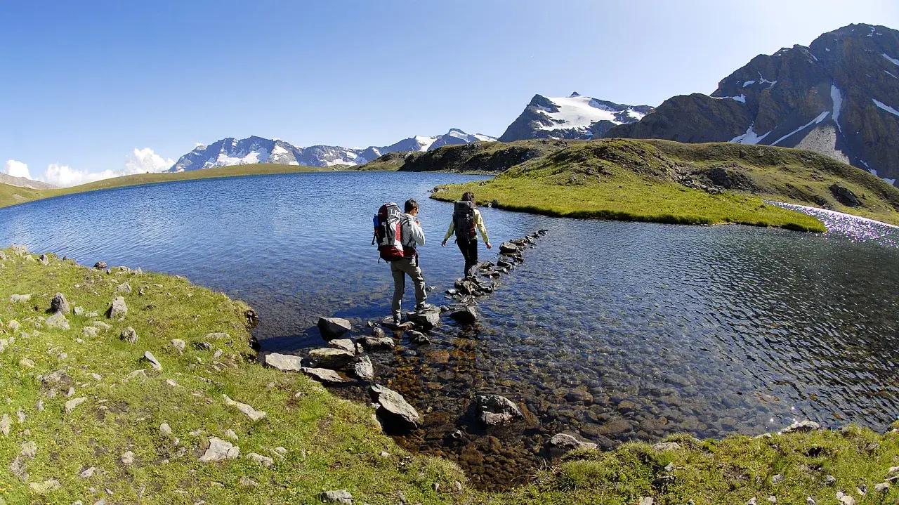 Due escursionisti attraversano il lago di Ceresole su un sentiero di pietre, con montagne innevate sullo sfondo.