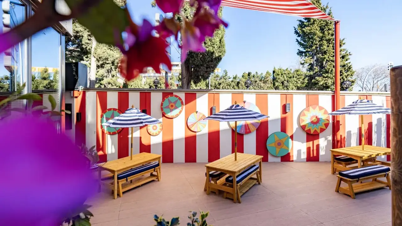Terraza de restaurantes con zona infantil en Madrid, decorada con rayas rojas y blancas y coloridos adornos circulares. Mesas de madera con sombrillas a rayas invitan a disfrutar.