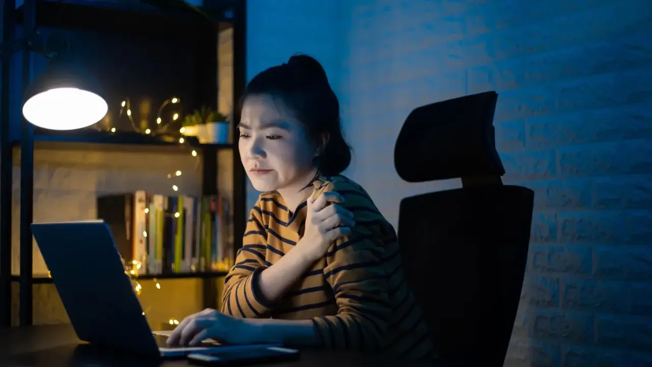 Une jeune femme, en pleine **mise en place télétravail**, ressent une douleur à l'épaule devant son ordinateur, éclairée par une lampe de bureau.