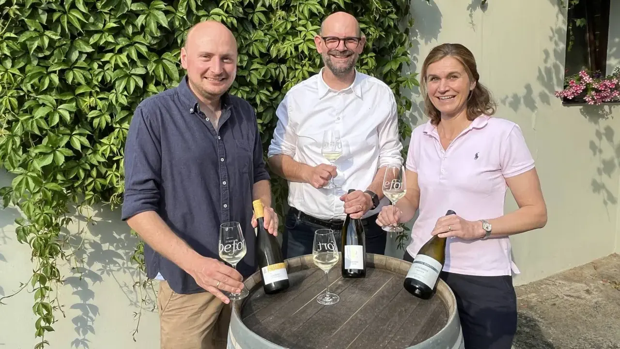 Drei Personen präsentieren Wein. Die Flasche mit Auxerrois Aussprache ist im Fokus.