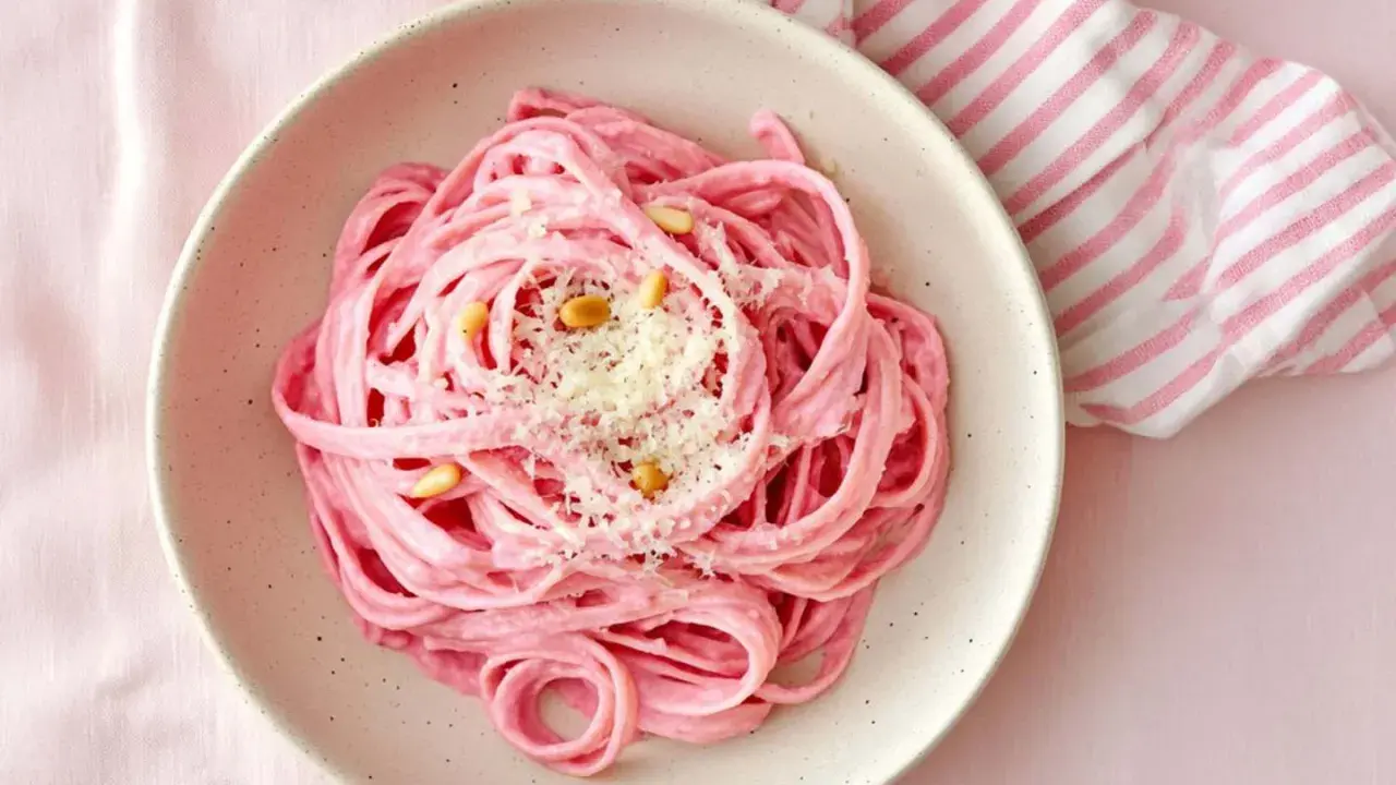 En skål med rosa pasta, toppad med pinjenötter och riven ost. En randig duk i rosa och vitt ligger bredvid.