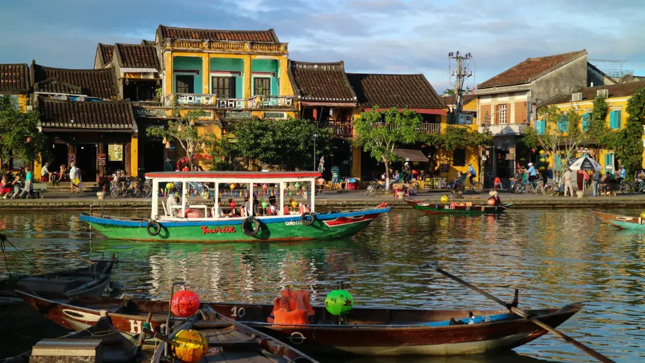 Båtar flyter på floden i Hoi An, Vietnam, med färgglada hus och folk som promenerar längs kajen.