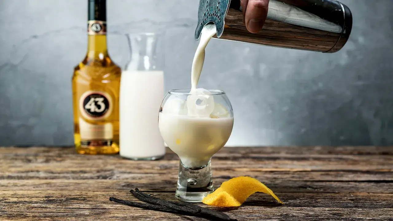 En hand häller en krämig dryck från en shaker i ett glas med is. Bredvid står en flaska Licor 43, en karaff med mjölk, vaniljstänger och citronskal.