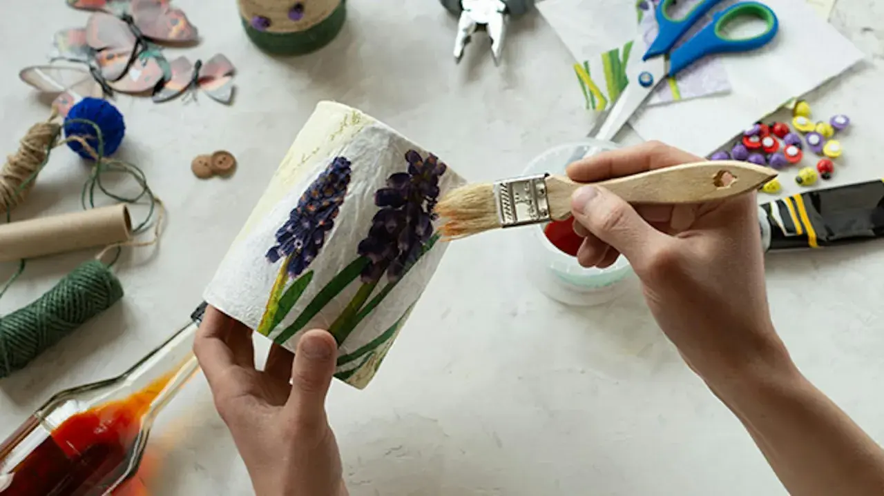 Ręce malują pędzlem ozdobiony kwiatami pojemnik, wokół materiały do decoupage.