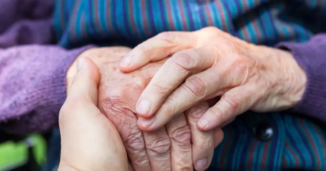 Dłonie młodszej osoby obejmują dłoń starszej, symbolizując wsparcie w obliczu choroby Alzheimera i jej przyczyn.