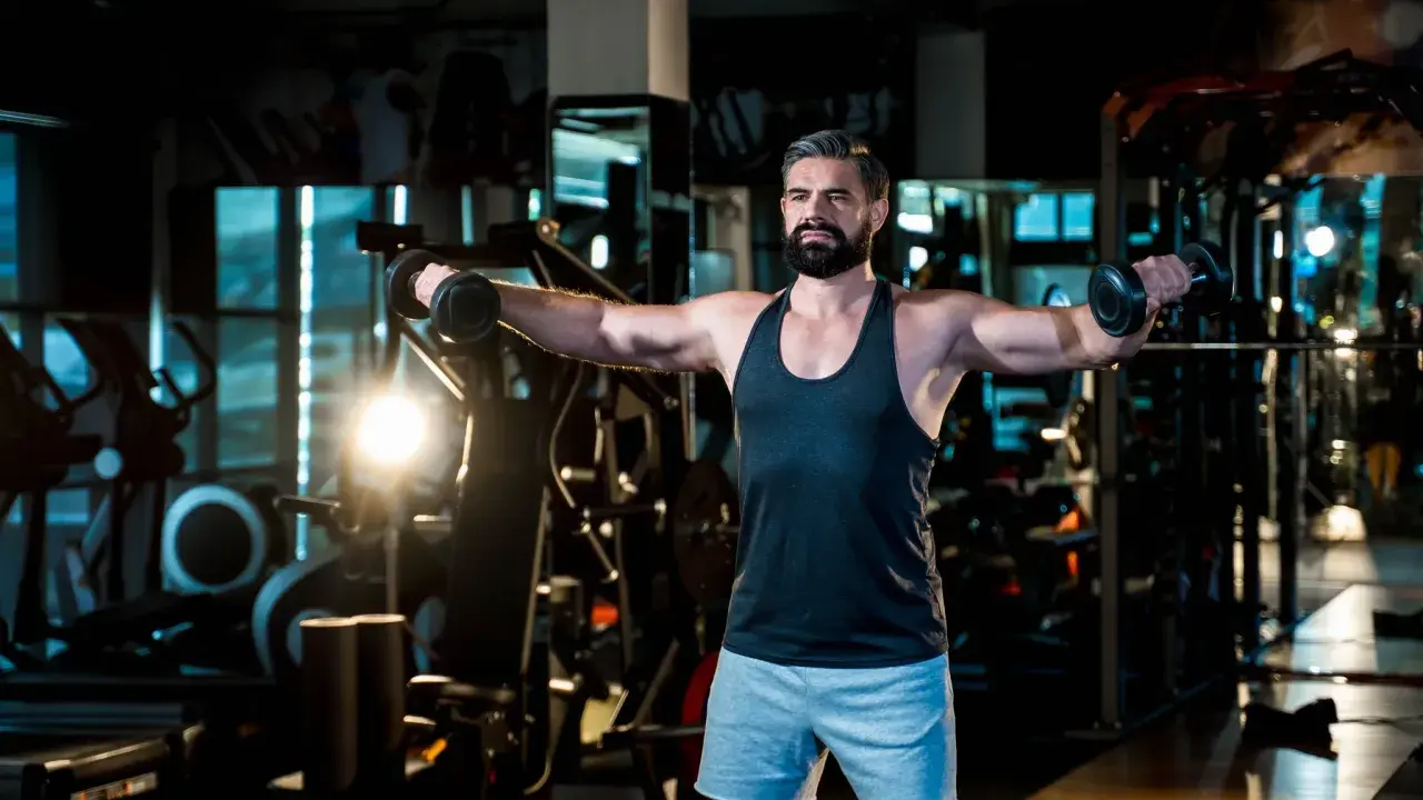 Mann mit Bart trainiert im Fitnessstudio, macht seitheben sitzend mit Hanteln.
