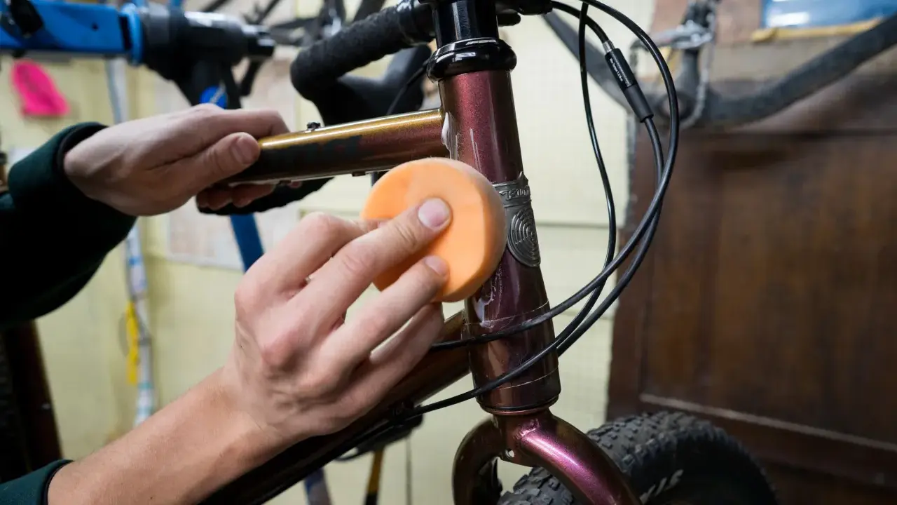 Hände polieren den Fahrradrahmen mit einem orangen Schwamm, um den Fahrradlack auszubessern.