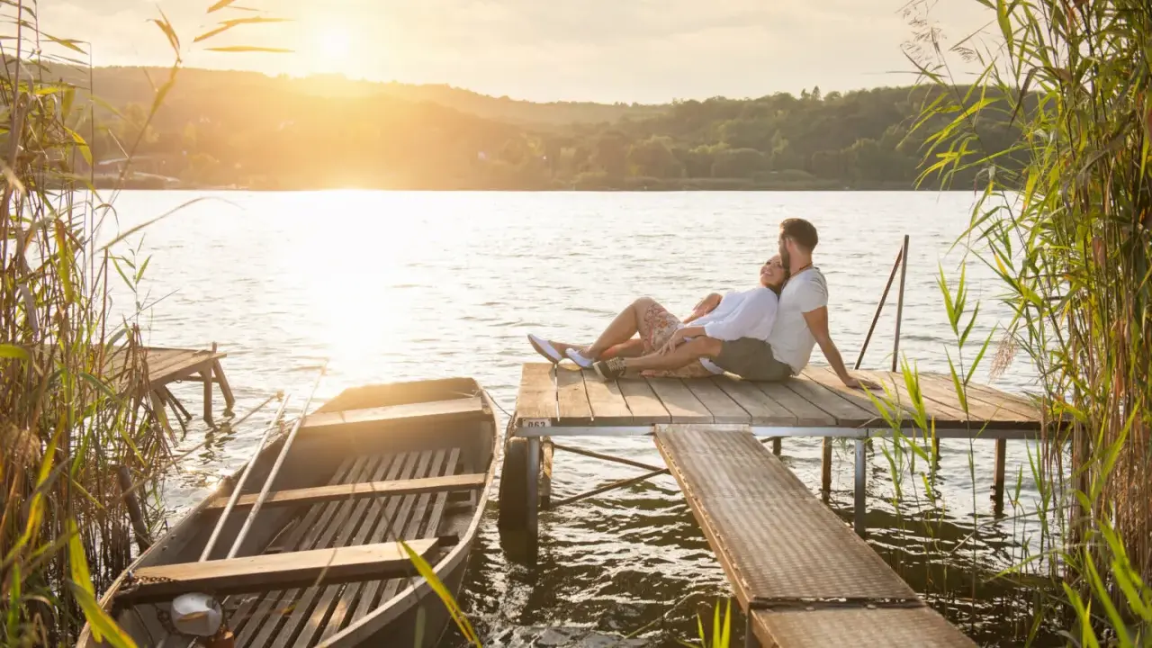 Para, gdzie na Mazury we dwoje? Para siedzi na pomoście, łódka przy brzegu, zachód słońca.