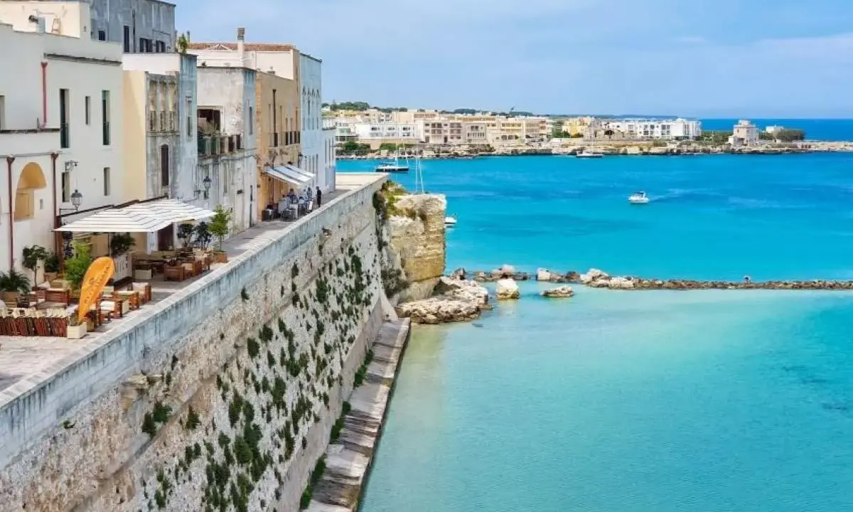 Vista del lungomare di Otranto centro, con edifici bianchi, un ristorante all'aperto e il mare azzurro cristallino.