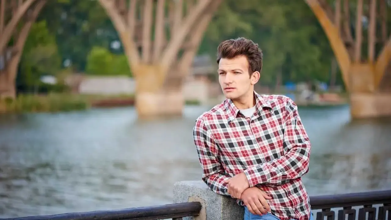 Młody mężczyzna w kratowanej koszuli i dżinsach opiera się o balustradę nad rzeką. W tle widać most.