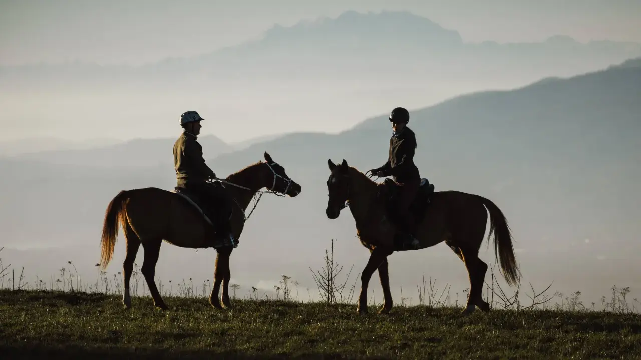 Due cavalieri in silhouette godono di una **passeggiata a cavallo in Lombardia** al tramonto, con montagne nebbiose all'orizzonte.