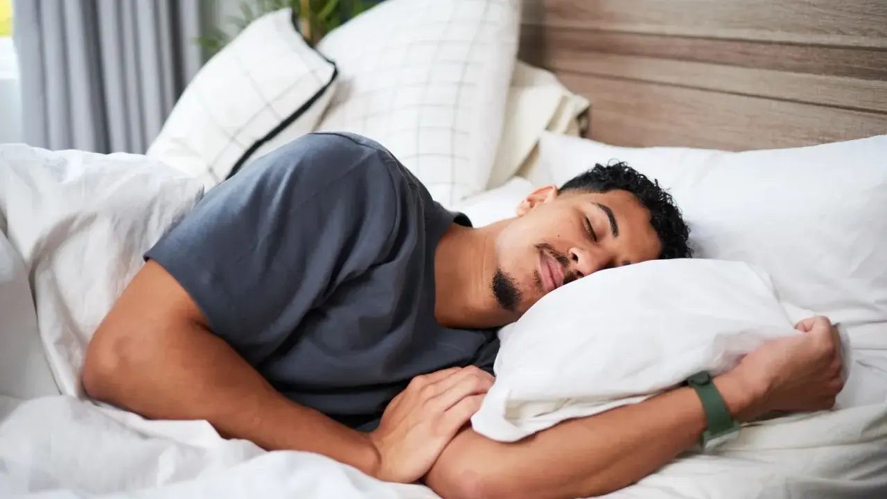 Ein Mann schläft tief und fest, um sein HRV zu verbessern. Er liegt auf einem weißen Bett mit vielen Kissen.