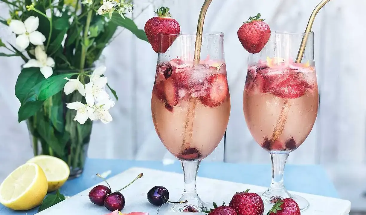 Två glas med drink med gin och prosecco, fyllda med is, jordgubbar och körsbär. Garnerade med jordgubbar och serverade med blommor och citron.