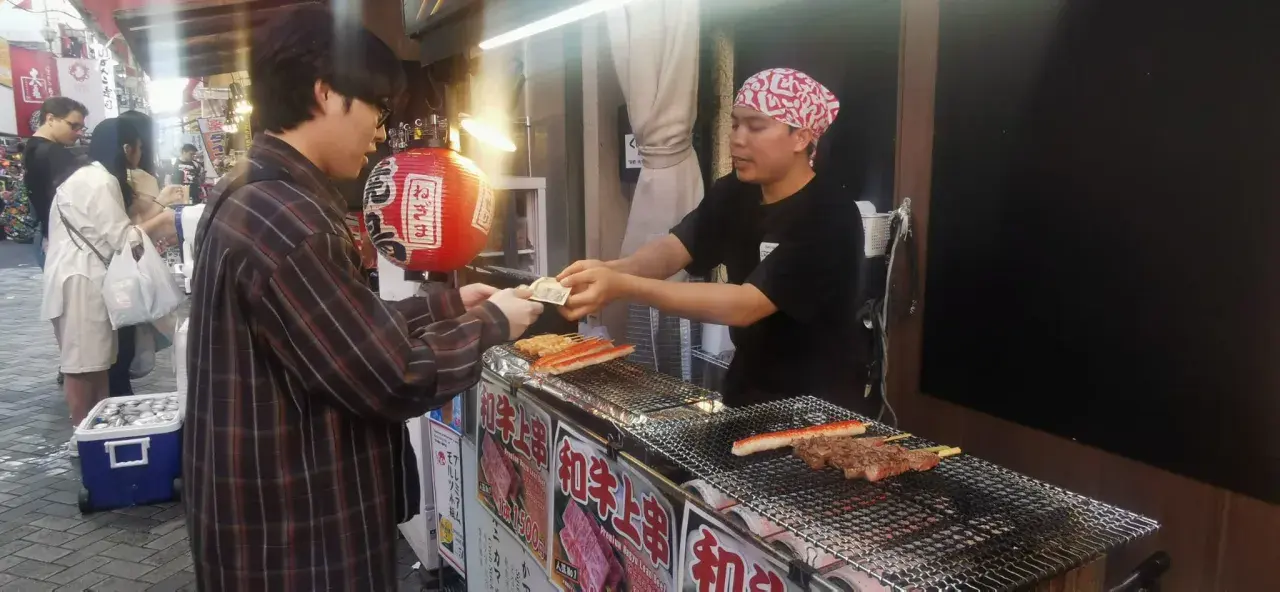 Un cliente paga por comida callejera japonesa: brochetas de carne y surimi a la parrilla en un puesto.