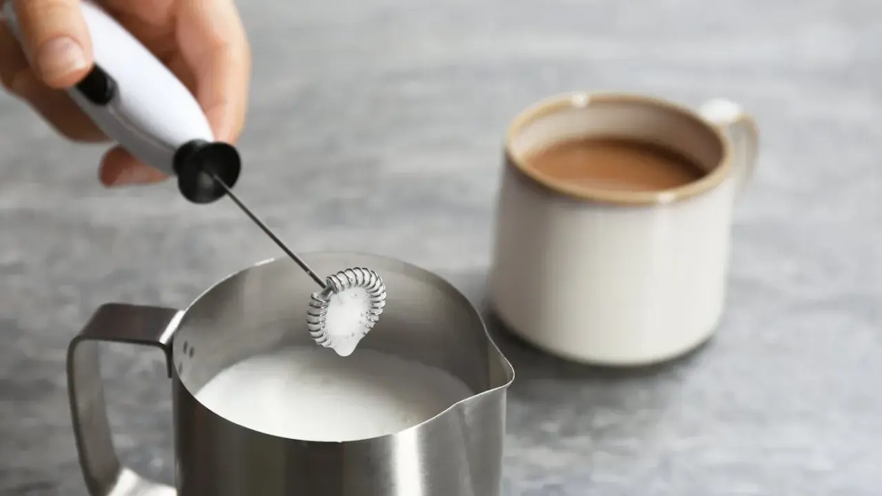 Hand hält Milchaufschäumer über Metallkännchen mit Milch. Perfekt für leckere Milchschaum Rezepte.