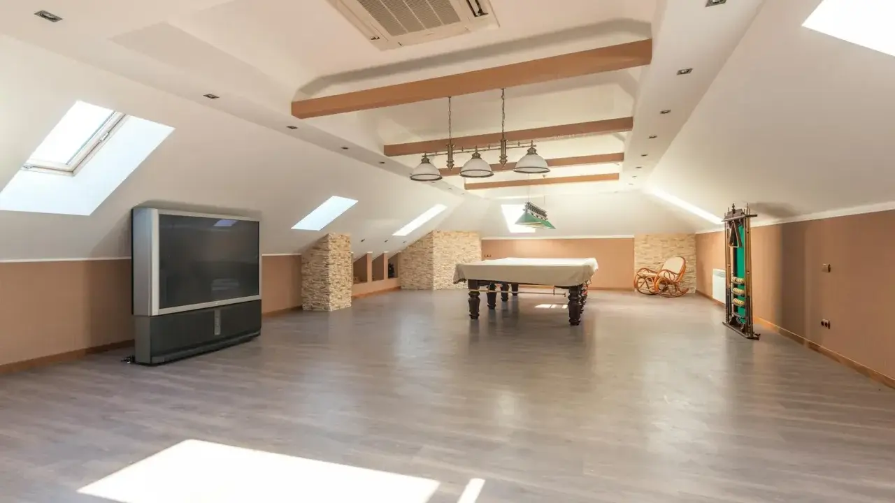 Vaste pièce sous les combles aménageables, avec table de billard, TV, fauteuil à bascule et étagères. Idéal pour un espace de loisirs.