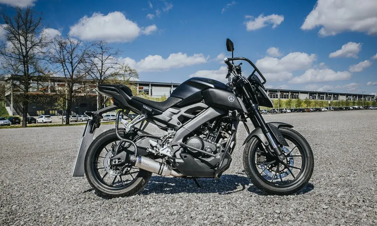 Czarny motocykl Yamaha MT-125, model kat a1, zaparkowany na żwirowym placu przed budynkiem.