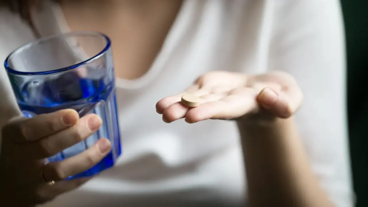 Une main tient un comprimé, peut-être un supplément de sels minéraux, à côté d'un verre d'eau bleue.