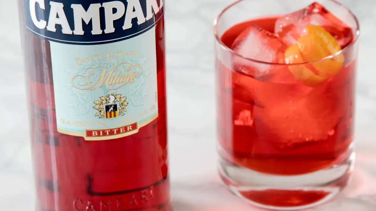 Butelka Campari i szklanka z czerwonym drinkiem, lodem i skórką pomarańczy. Idealny aperitif, który może być bazą dla drinków z Aperolem.