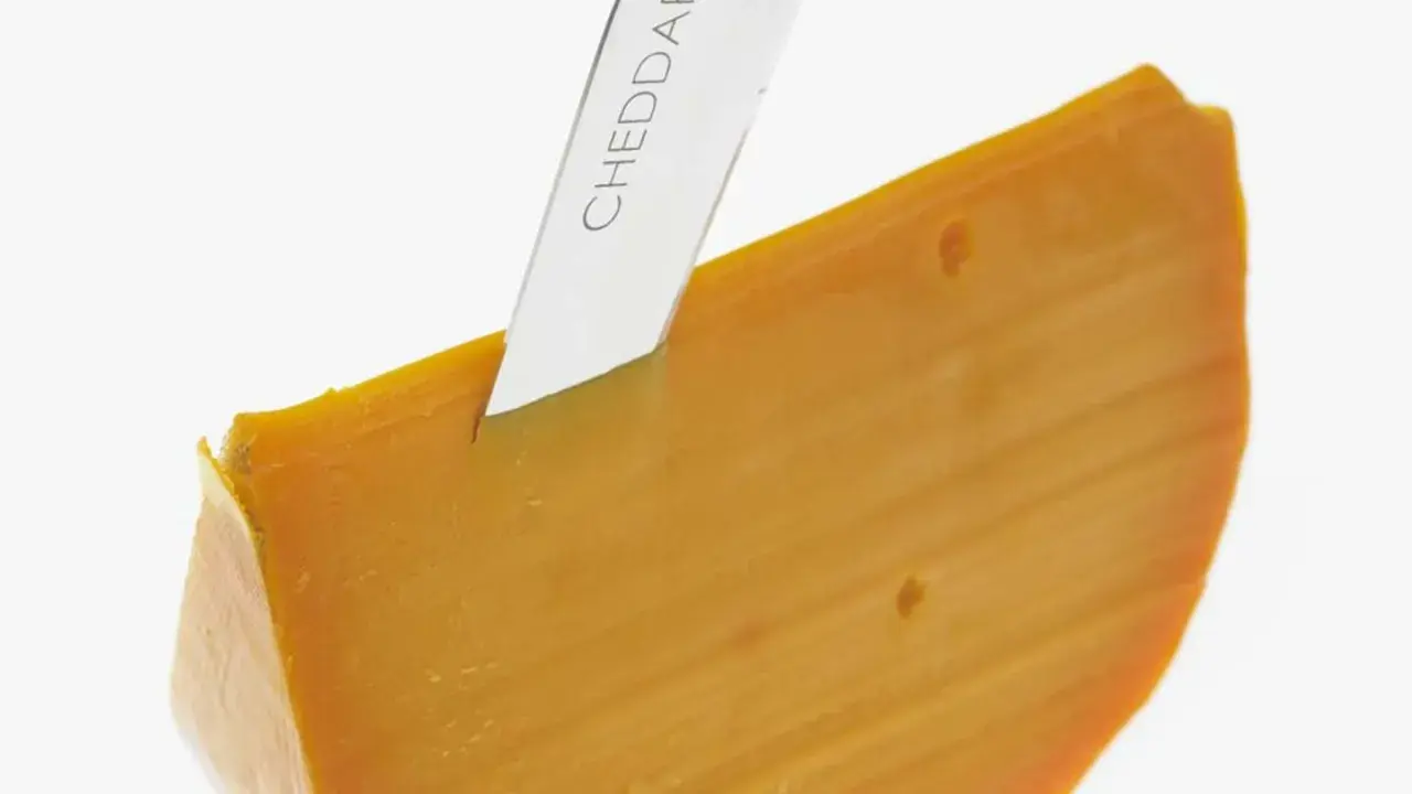 Ein Messer schneidet ein Stück leuchtend orangefarbenen Cheddar-Käse. Was ist Cheddar? Ein köstlicher Hartkäse.
