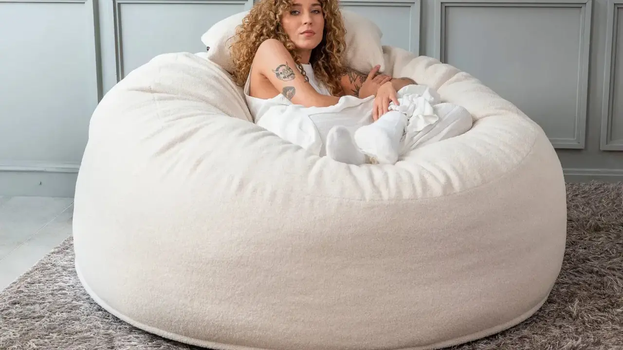 En kvinna med lockigt hår vilar i en stor, mjuk beige sittpall.