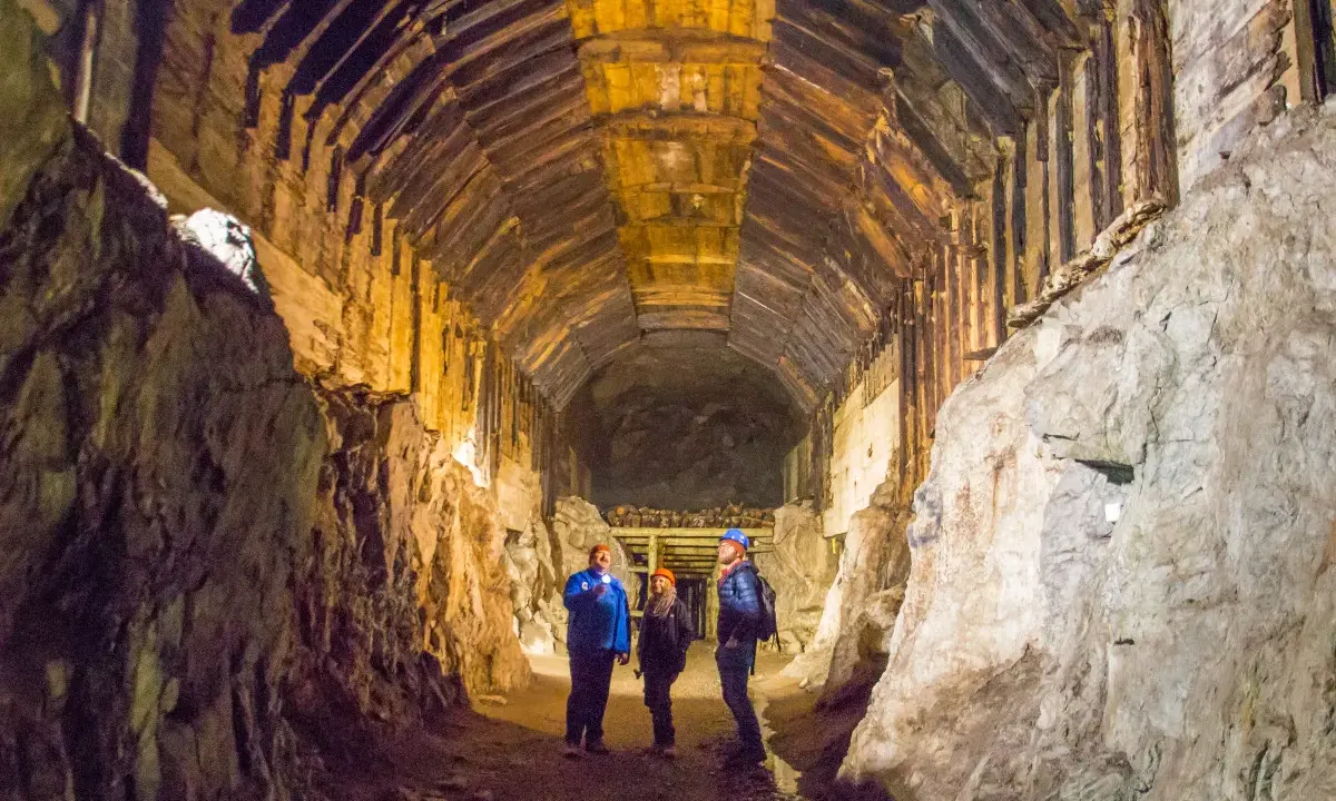 Trzech turystów w kaskach eksploruje stary tunel kopalni z drewnianymi wzmocnieniami i skalnymi ścianami.