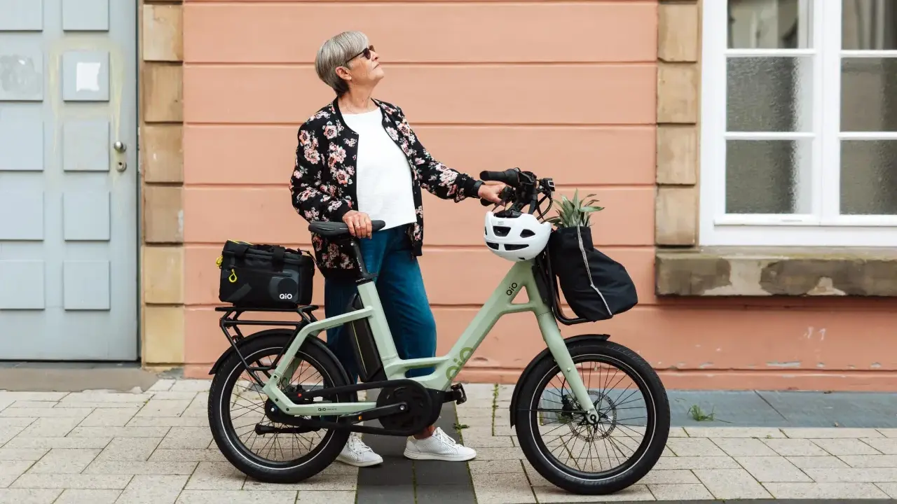 Ältere Dame mit QIO Bike, positive Erfahrungen mit dem E-Bike, das mit einem Helm und einer Tasche beladen ist.