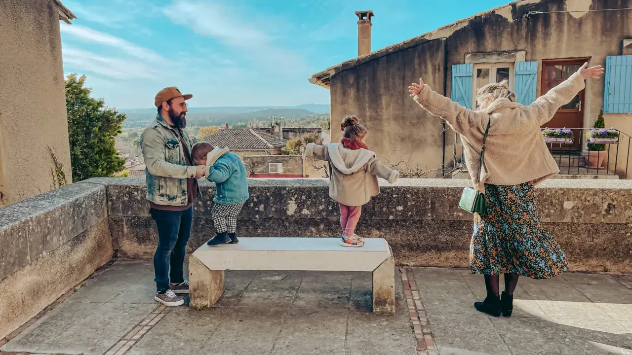 Famille profitant d'une vue panoramique depuis un balcon à Châteauneuf de Gadagne. Le père tient un enfant, tandis que deux autres enfants jouent sur un banc.