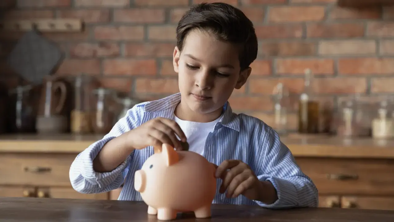 Niño introduce una moneda en una hucha rosa, aprendiendo sobre el ahorro con la cuenta infantil BBVA.