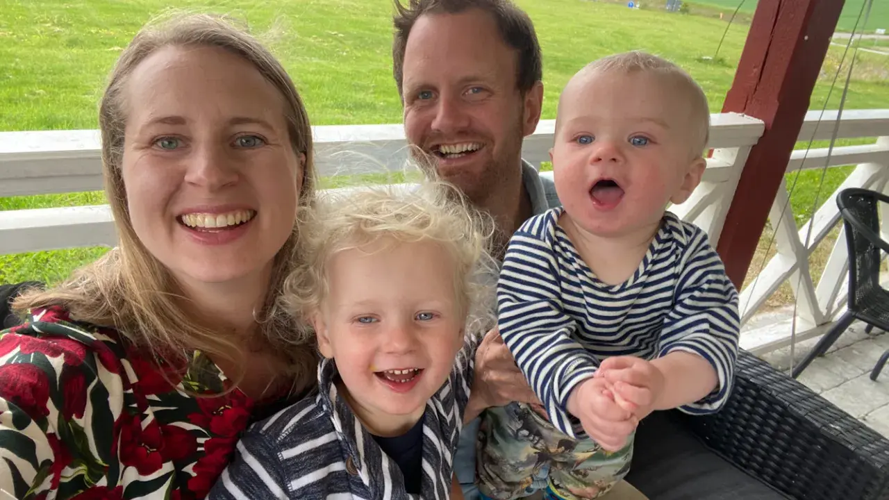 Glad familj njuter av utomhusaktiviteter. Två små barn och deras föräldrar ler mot kameran på en veranda med grönskande fält i bakgrunden.