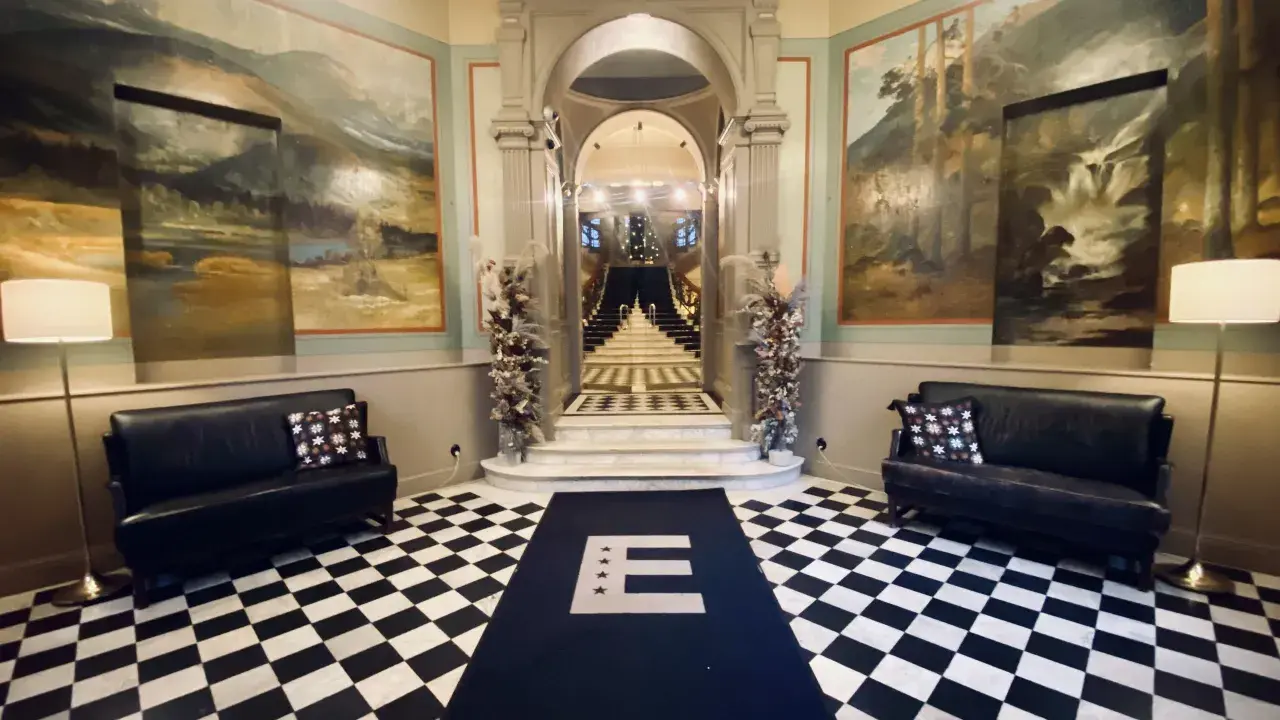 Elegant foajé med schackrutigt golv, konstverk på väggarna och en trappa. En matta med bokstaven E leder mot trappan. Känns som en plats med mycket hotell Knaust historia.