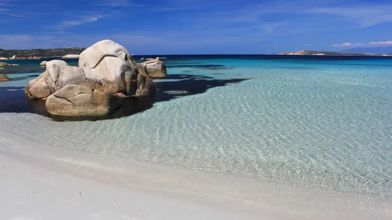 Spiaggia bianca e rocce granitiche sull'isola della Maddalena, con acque cristalline e un cielo azzurro intenso.